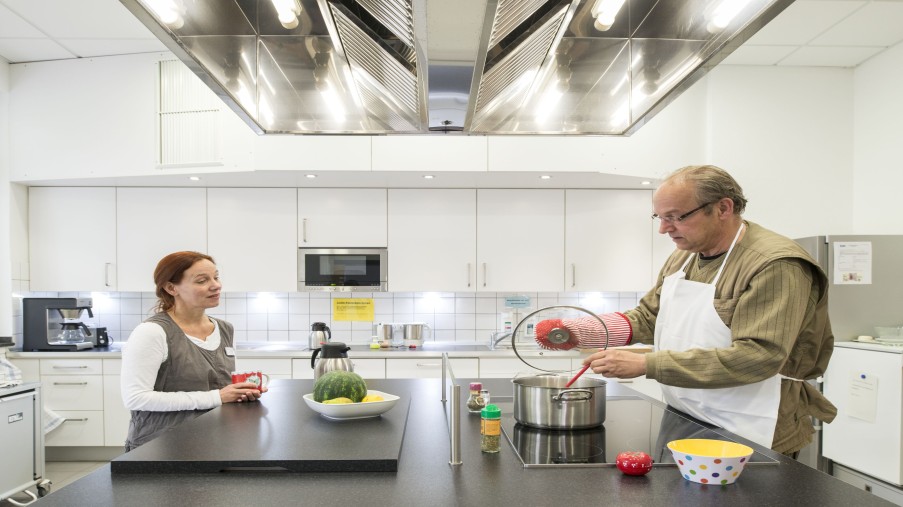 Mann kocht in einer großen Stationsküche, Frau schaut ihm - eine Kaffeetasse haltend - dabei zu.