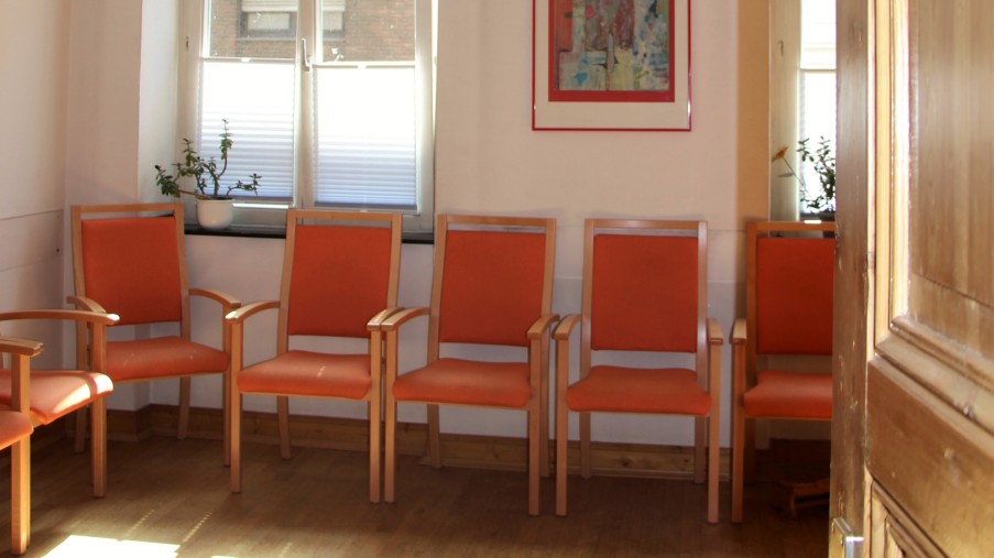 Ein Therapieraum mit roten Stühlen, hohen Decken und einer stuckverzierten Decke.