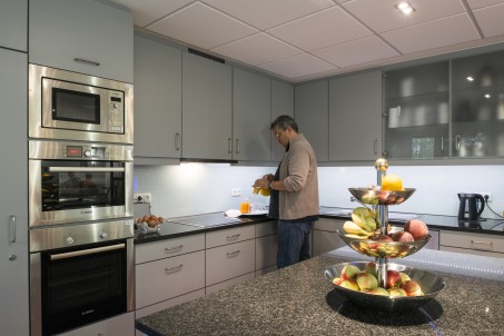 Das Bild zeigt eine geräumige Küche mit modernen Elektrogeräten. Im Vordergrund ist eine Etagere mit Obst zu sehen. Im Hintergrund bereitet ein Mann ein Gericht zu.